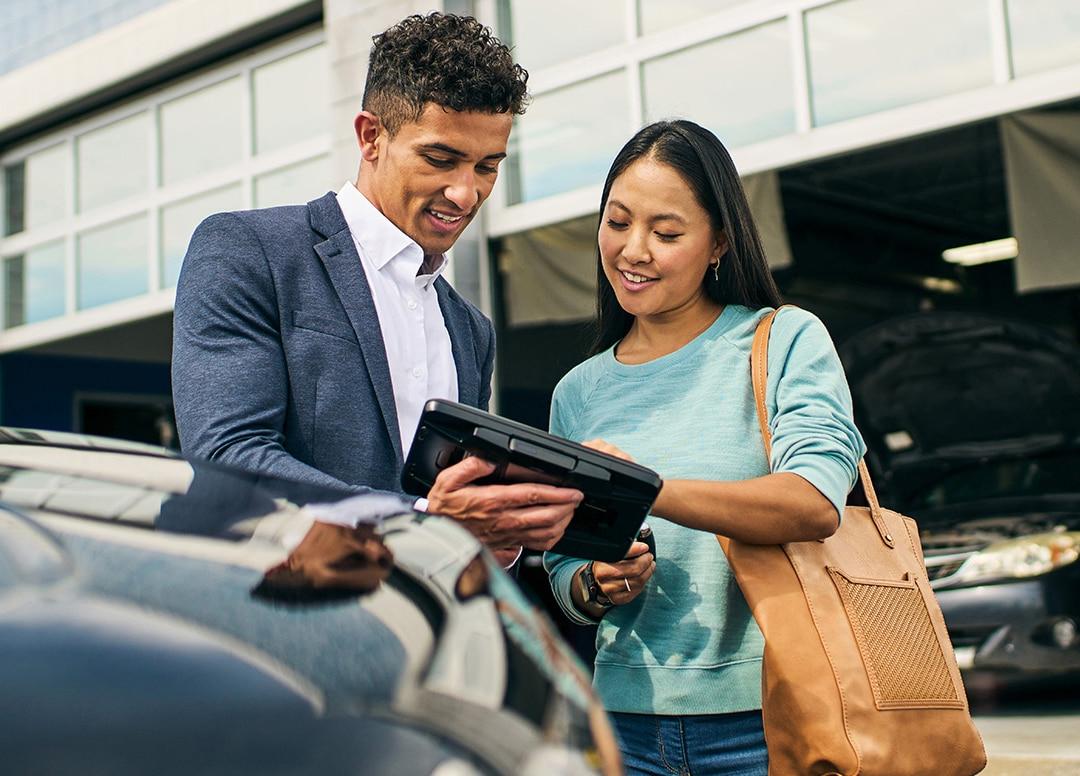 一名棕色肤色的女性企业员工拿着手机，陪着一名浅色肤色的男性顾客，带着一个手提箱走出企业停车场.